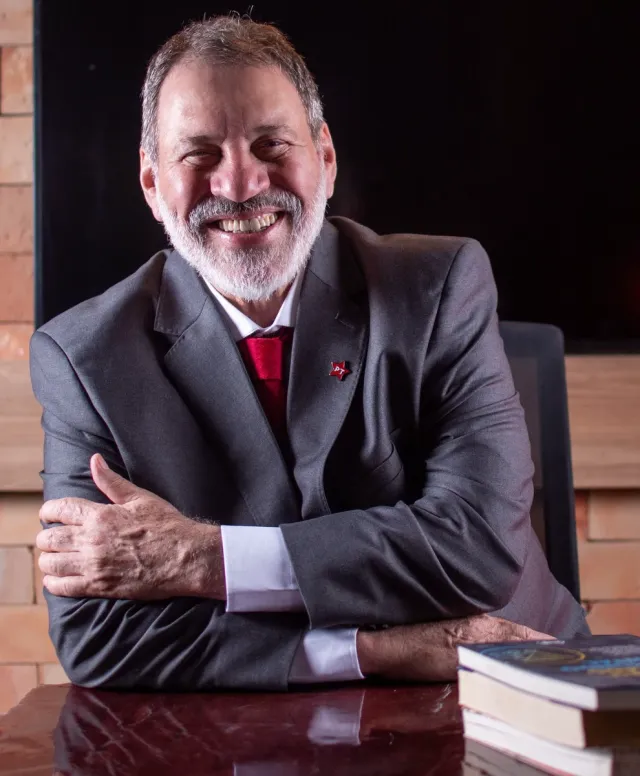 Em foto, Delúbio Soares, homem branco e grisálio, sorri para foto. Esta vestido com um terno cinza, gravata vermelha e um broche do PT na gola esquerda.