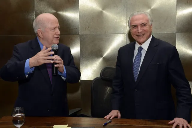 Albano Franco que tomou posse na Assembleia Legislativa de Sergipe e Michel Temer, dois homens de terno em um ambiente formal; um deles fala ao microfone enquanto gesticula, e o outro sorri ao lado de uma mesa com um copo de água.