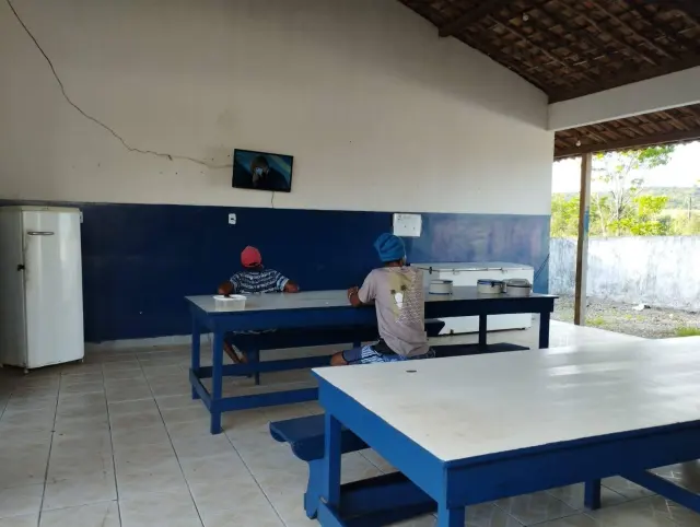Refeitório simples com mesas e bancos azuis; duas pessoas sentadas de costas assistem a uma televisão pequena fixada na parede, nas plantações de cana em Sergipe