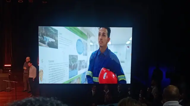 Apresentação em audiência pública mostra no telão um trabalhador da empresa Loga, de capacete de segurança, enquanto palestrantes falam ao público sobre projeto de incinerador de lixo na cidade.