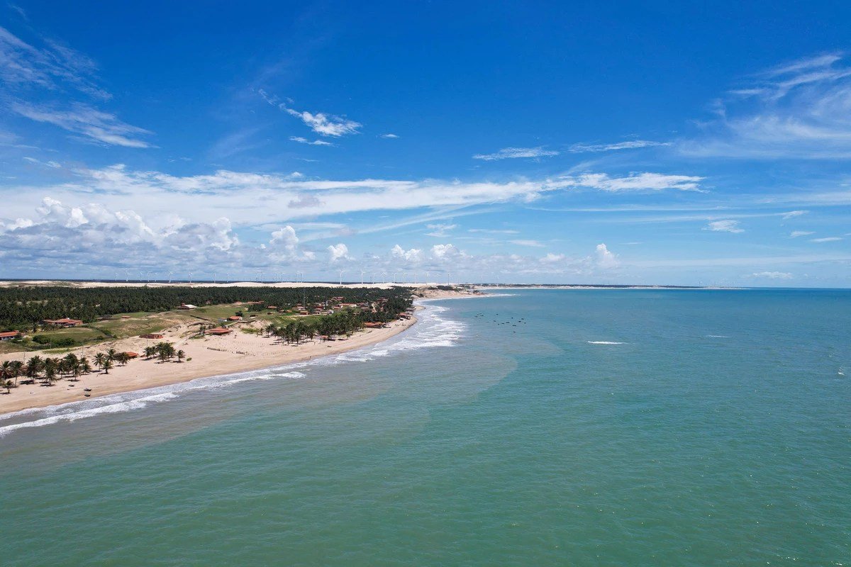 Imagem aérea da costa cearense mostra divisa entre mar e faixa de areia, com turbinas eólicas ao horizonte