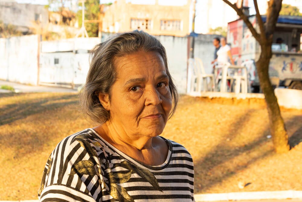 Maria Lúcia, moradora de São Miguel Paulista. Maria é uma senhora na faixa dos 60 anos, com cabelos grisalhos e pele parda. Ela está em uma praça pública e veste uma camiseta preta e branca listrada com o desenho de uma flor no ombro esquerdo