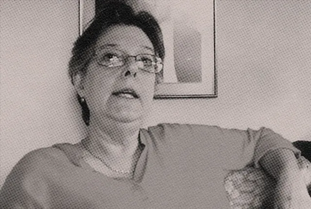 Lygia Jobim é uma mulher na faixa dos 70 anos, de cabelos grisalhos e óculos. Ela está sentada em um sofá